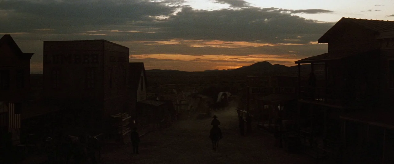 Tombstone (1993) movie still: establishing — A quiet Old West town street with wooden facades lines a dusty central path. A single rider on…
