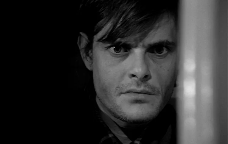 Werckmeister Harmonies (2000) movie still: closeup — A man looks through a sliver of an opening in a dark room. Deep shadows obscure the rest of the…