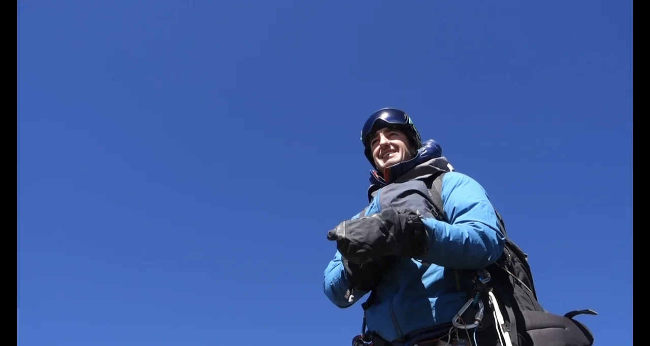 Blutch (2018) movie still: medium — A climber stands on a steep mountain slope against a clear deep blue sky. The man is wearing technical climbing…