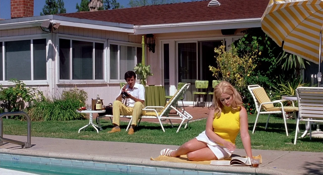 Viva (2007) movie still: wide — A suburban ranch-style house with a backyard pool area containing lawn chairs and furniture. A blonde woman sits…