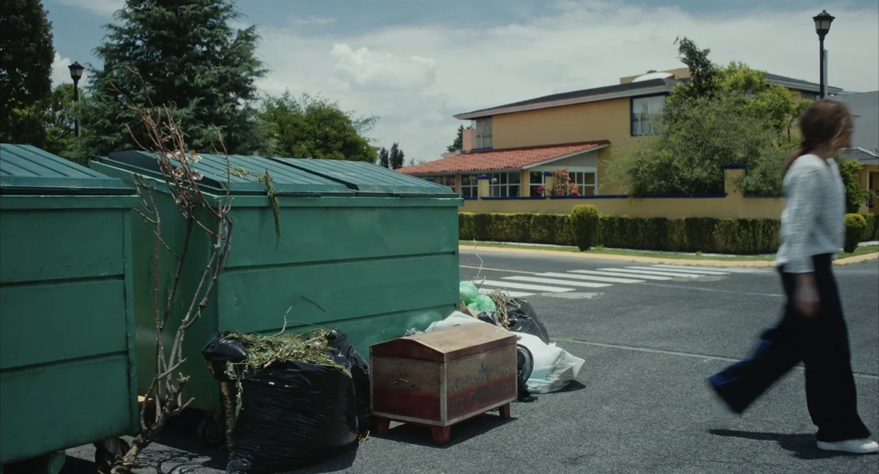 The Diary (1999) movie still: medium — A quiet suburban street features green industrial dumpsters parked beside a paved road. A woman in a light…