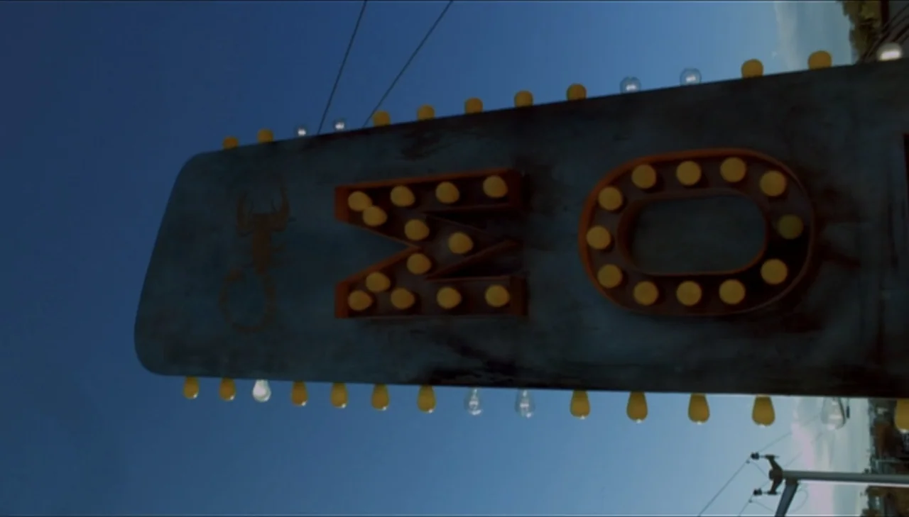 Dark Country (2009) movie still: closeup — A close-up view of a dark metal motel sign against a deep blue sky. The sign features a small metal…