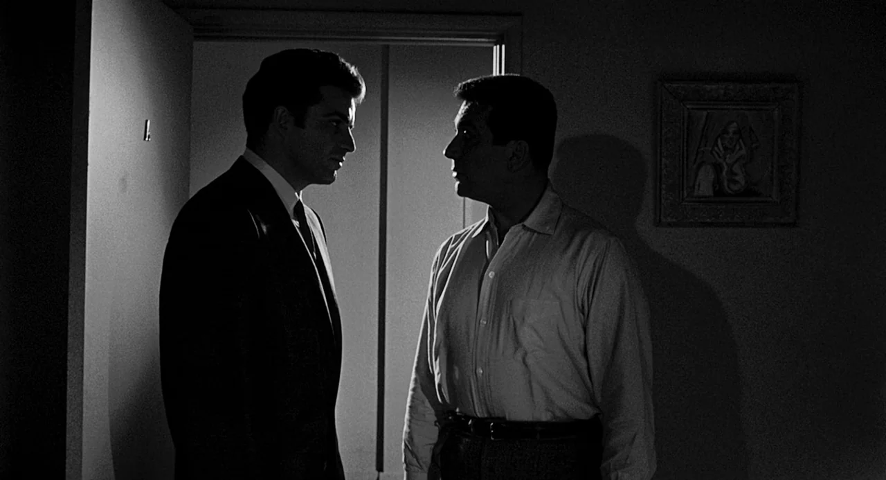 Murder By Contract (1958) movie still: medium — A dark hotel room interior features two men standing in front of a doorway. The figures are…