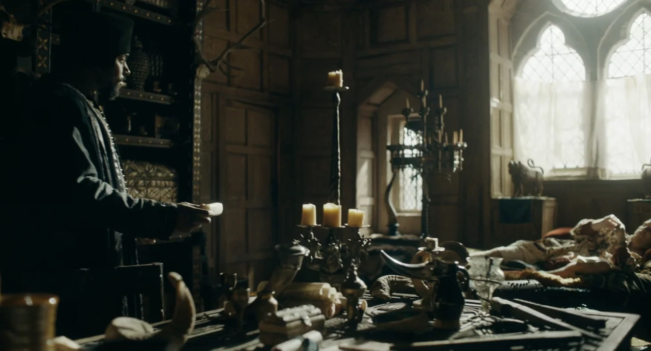 Catherine Called Birdy (2022) movie still: medium — A stone-walled medieval room features dark wood paneling and a large window with sunlight. A man…