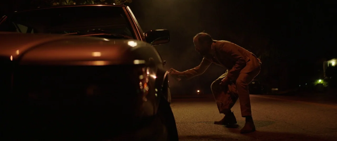 Lemon (2017) movie still: medium — A man in a suit crouches beside a dark sedan on a dimly lit street. He holds a spray can near the vehicle's rear…