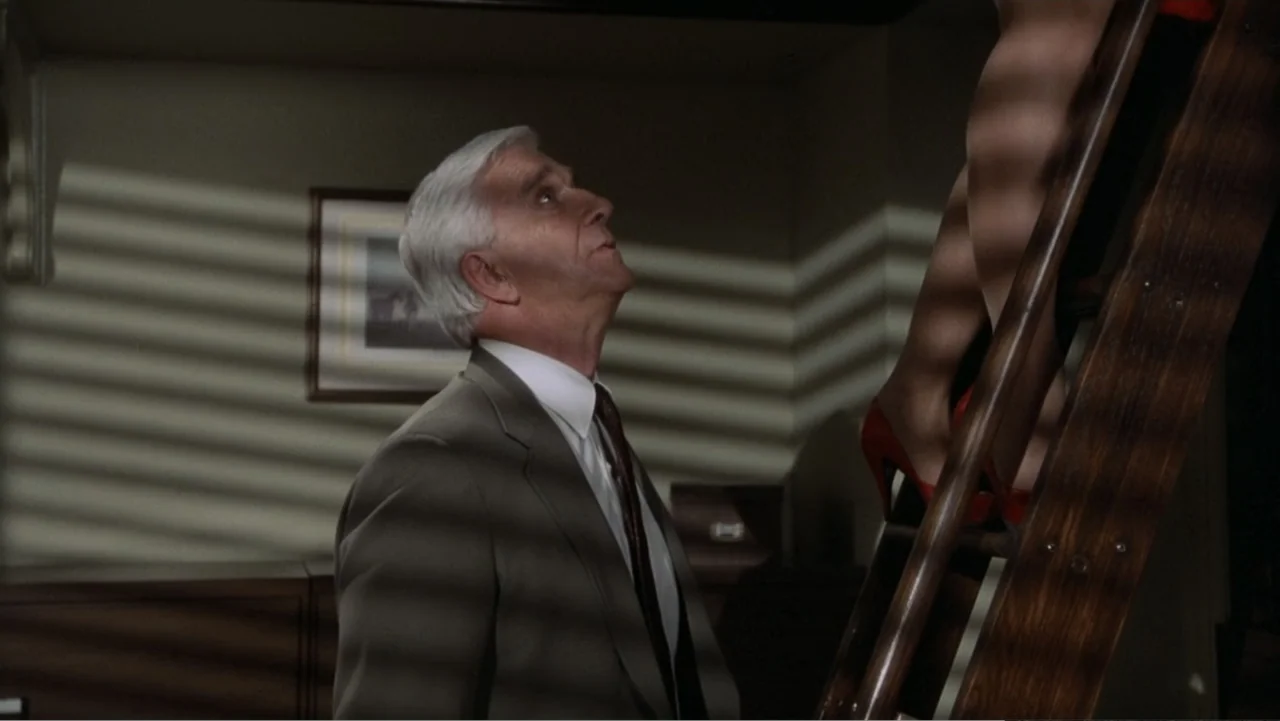 The Naked Gun: From the Files of Police Squad! (1988) movie still: medium — An office interior features a man standing near a wooden ladder holding…