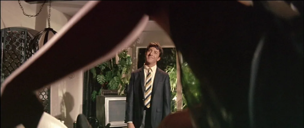 The Graduate (1967) movie still: medium — A 1960s living room features a houseplant and a vintage television set. The male subject stands in the…