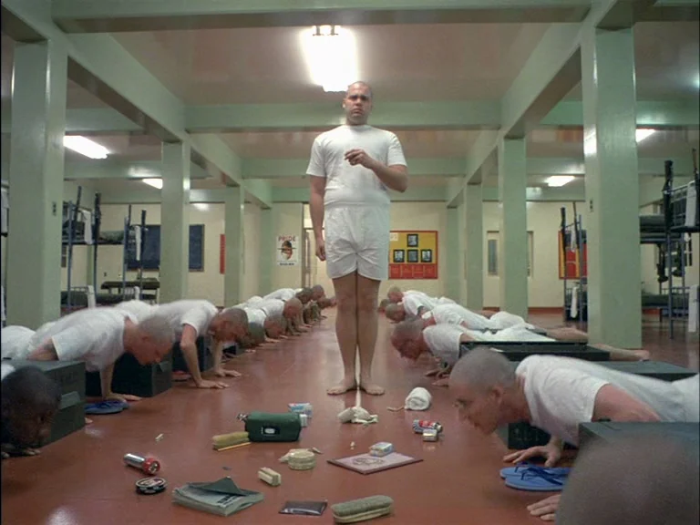 Full Metal Jacket (1987) movie still: fullbody — A military barracks hall features rows of bunk beds and concrete pillars painted mint green.…