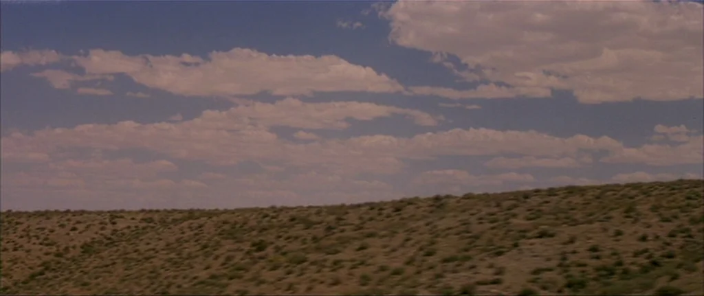 Gerry (2002) movie still: establishing — An arid, scrub-covered hillside stretches across the lower portion of the frame. A wide blue sky with…