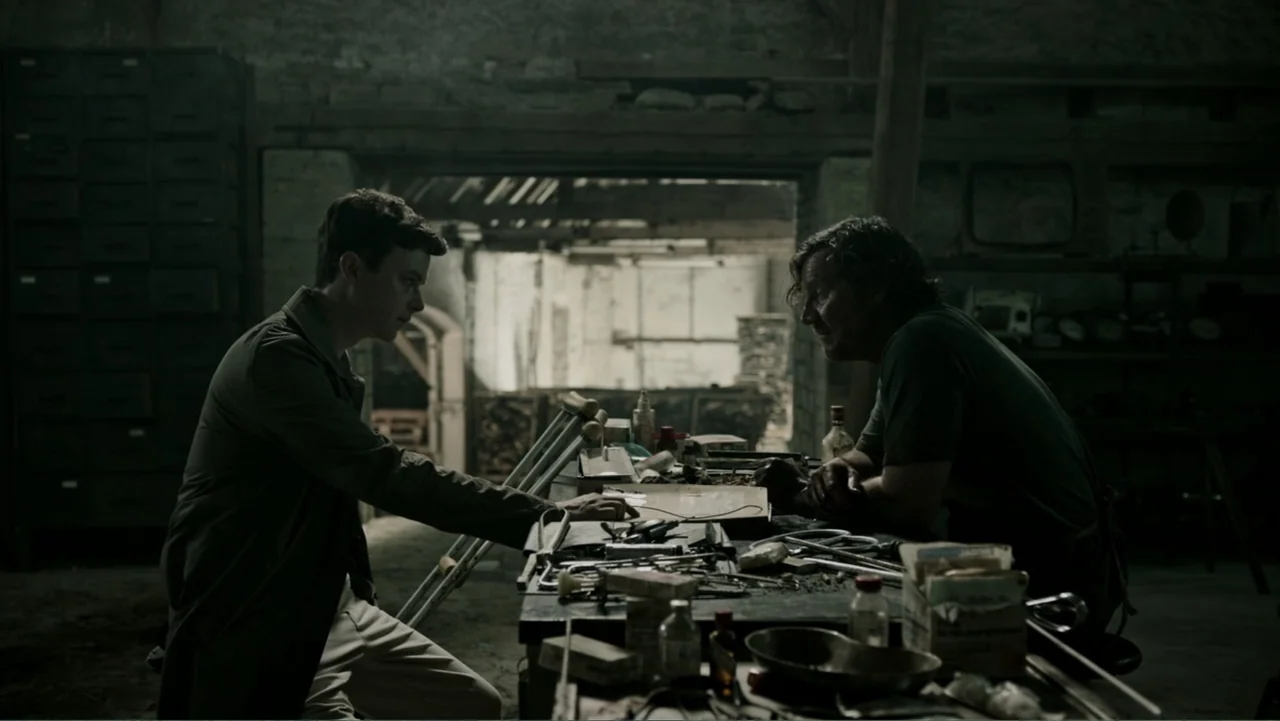 A Cure for Wellness (2017) movie still: medium — A cluttered, dimly lit workshop features a heavy table covered in various metal tools. Two men sit…