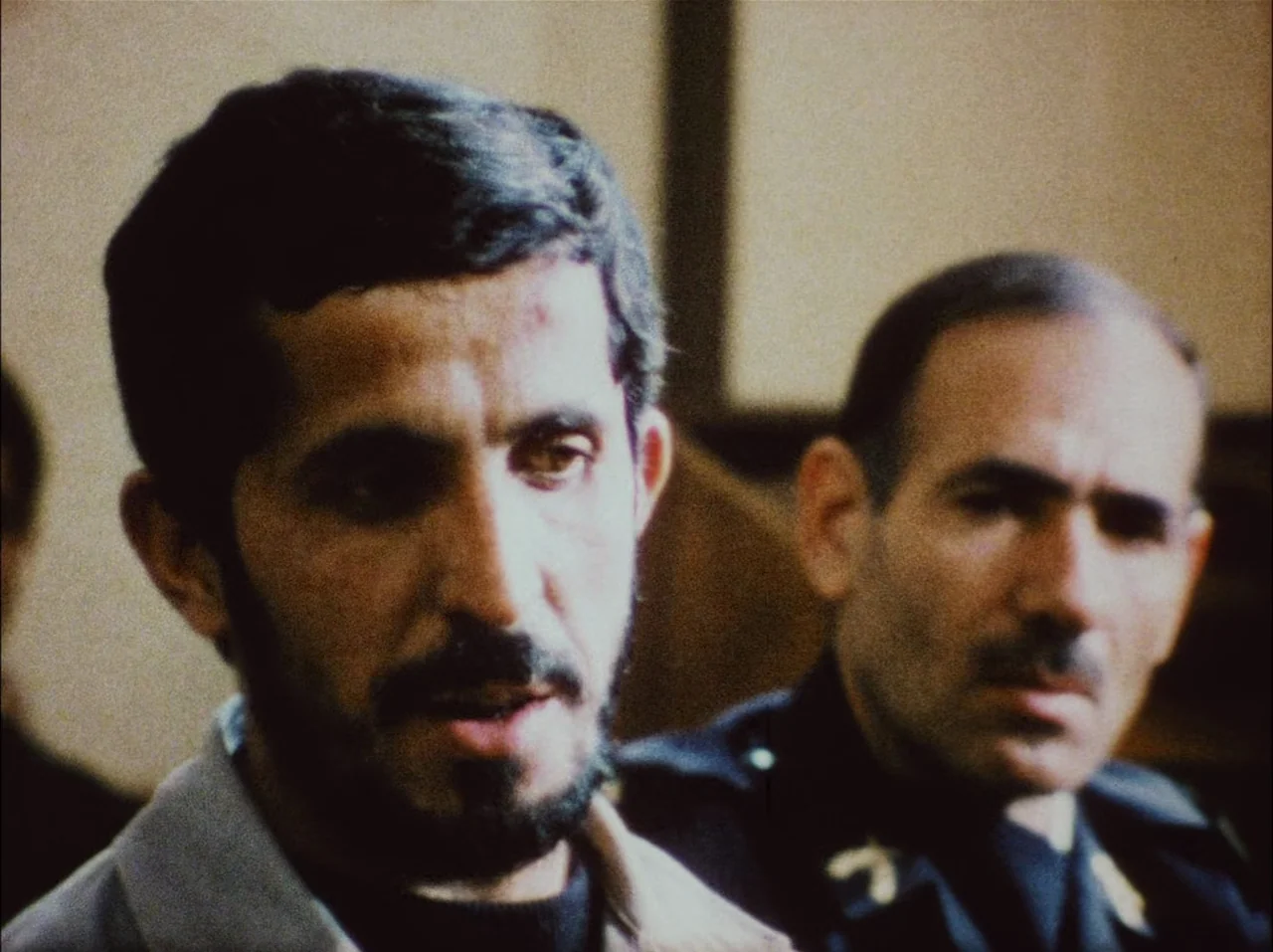 Close-Up (1990) movie still: medium — An indoor setting with indistinct beige walls suggesting an office or courtroom environment. A bearded man…