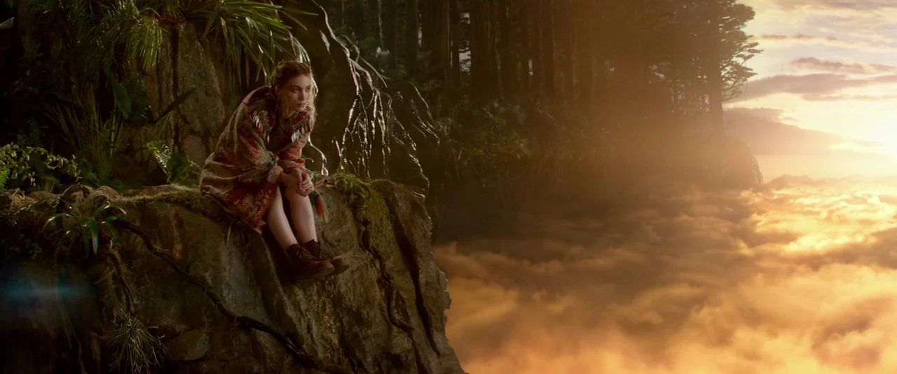 Pan (2015) movie still: wide — A woman sits on a rocky cliff edge high above a dense cloud layer. Dark trees grow behind her while the sun sets…