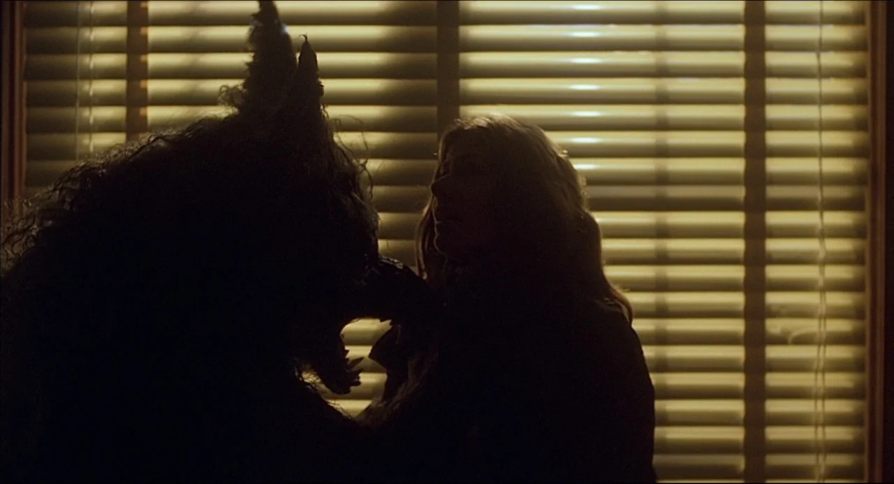 The Howling (1981) movie still: medium — The silhouettes of a woman and a creature stand before a window with blinds. The scene is illuminated only…