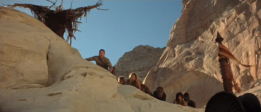 Planet of the Apes (2001) movie still: wide — A group of humans climbs a sun-bleached sandstone ridge in a vast rocky desert. A dark, skeletal-like…