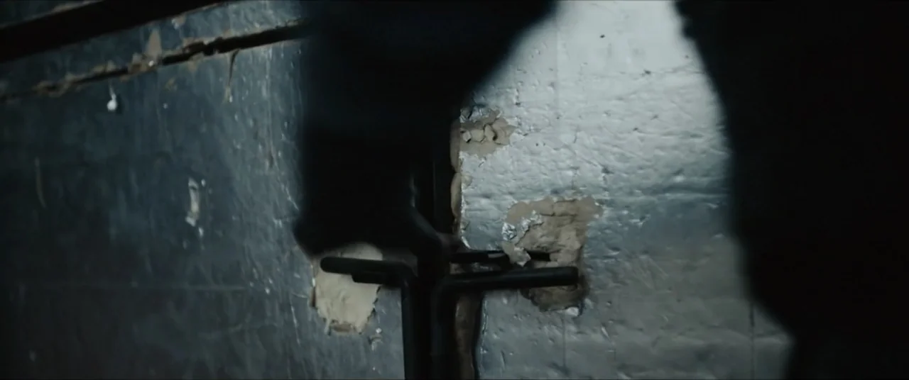Vicious Fun (2021) movie still: closeup — A textured silver-grey metallic wall features cracked plaster and dark vertical structural beams. A human…