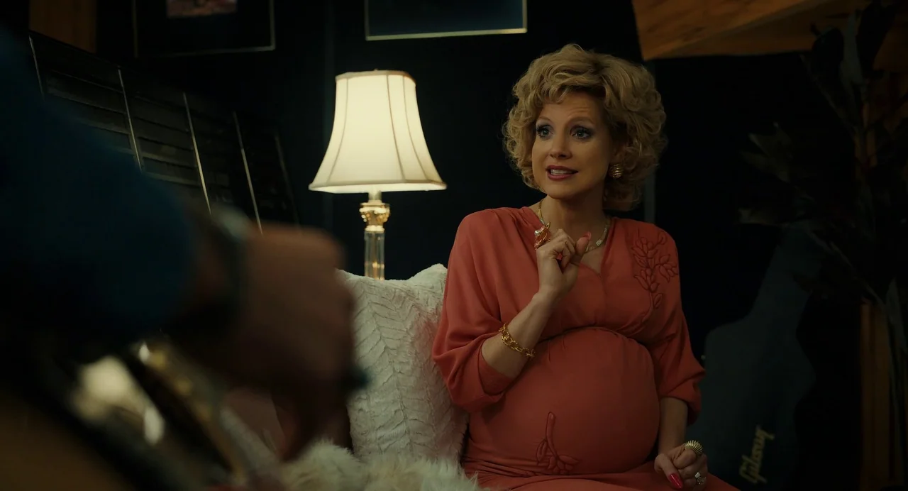 The Eyes of Tammy Faye (2021) movie still: medium — A dimly lit room with black walls and a prominent lamp behind a woman. The pregnant woman sits…