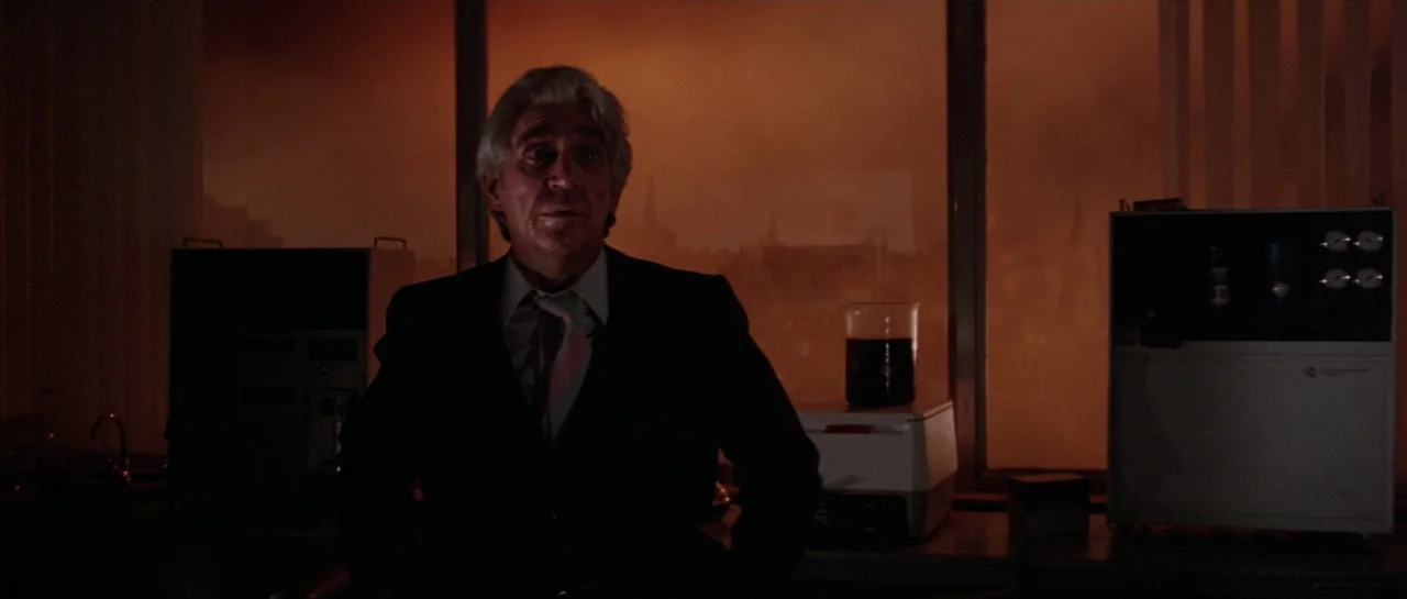 Lifeforce (1985) movie still: medium — A dark laboratory interior has a large window overlooking an orange, hazy exterior view. An older man in a…