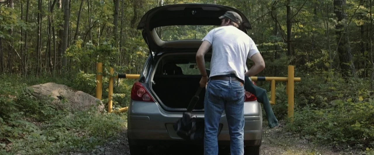 Darkness Waits (2020) movie still: medium — A silver hatchback sits on a dirt track in a dense deciduous forest. A man stands at the rear of the…
