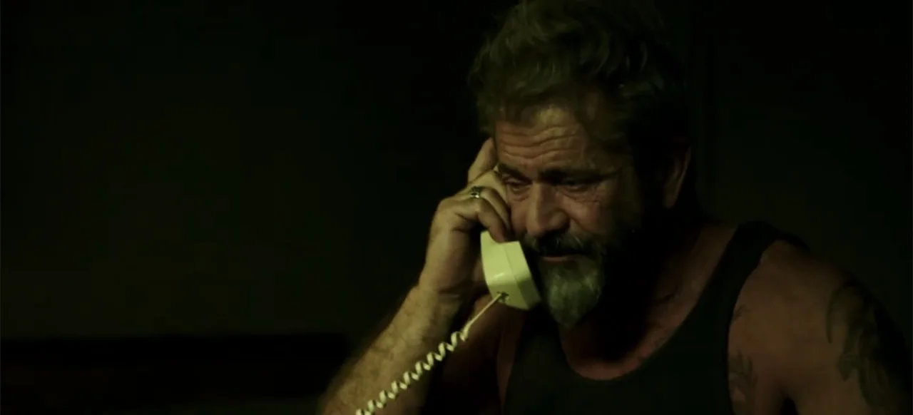 Blood Father (2016) movie still: closeup — A dimly lit room holds a man in a dark tank top. The man holds a telephone handset to his ear while…