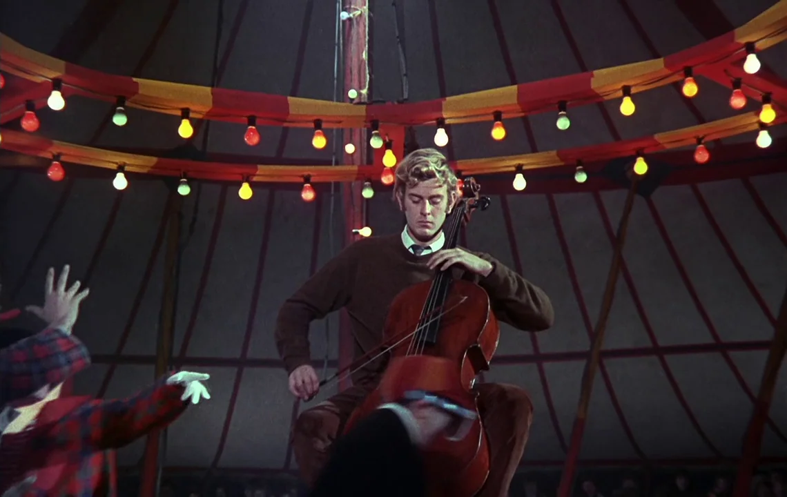 The Girl on a Motorcycle (1968) movie still: medium — The interior of a circus tent is decorated with circular strings of hanging bulbs. A young man…