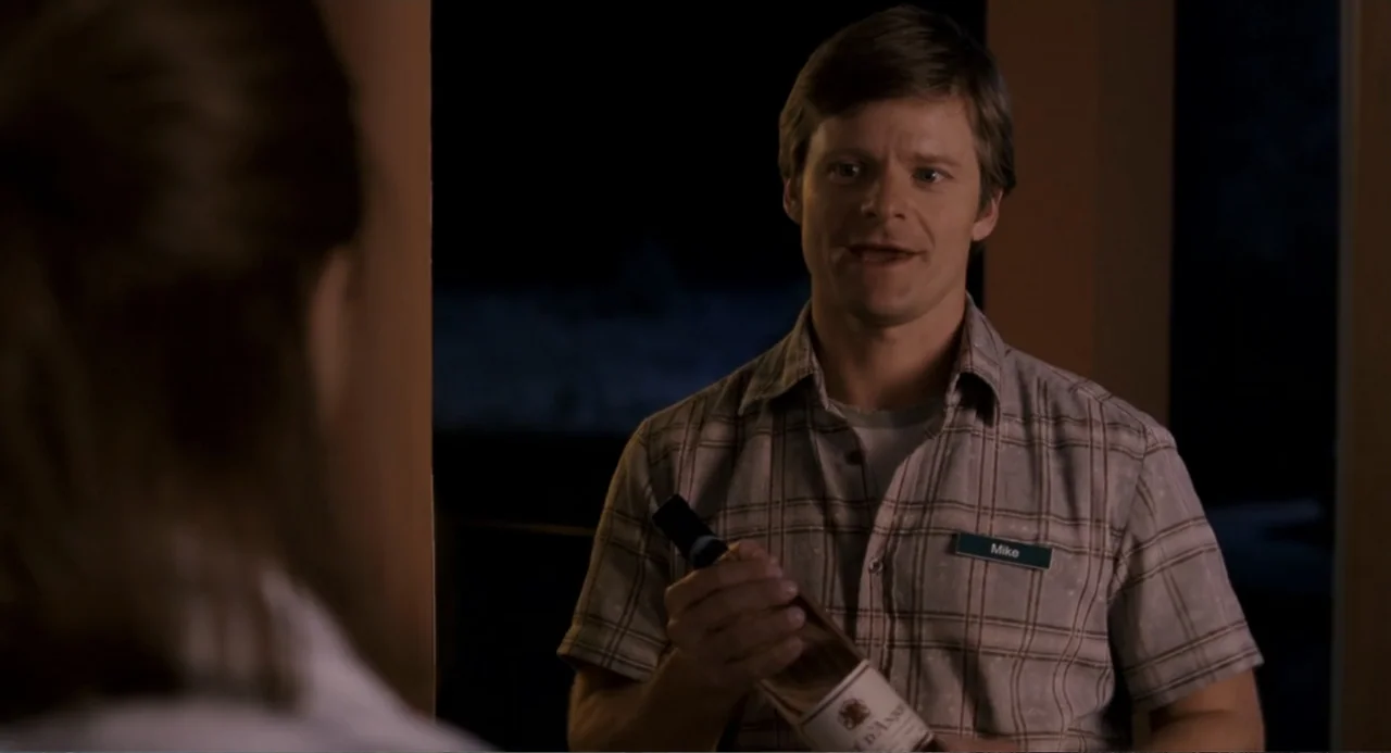 Management (2009) movie still: medium — A man stands indoors at night wearing a store employee name tag and uniform. He holds a bottle of wine in…