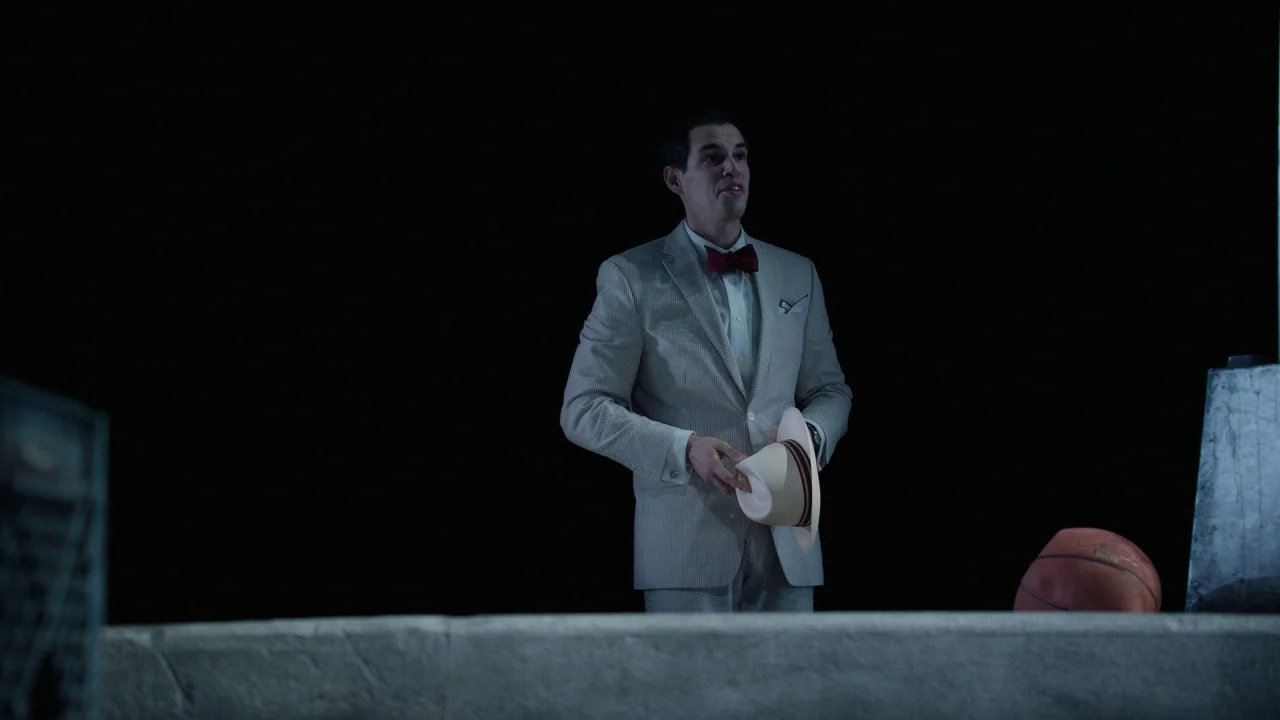 Pass Over (2018) movie still: medium — A man stands on a dark, flat surface against an entirely black void. He is holding a hat while looking…