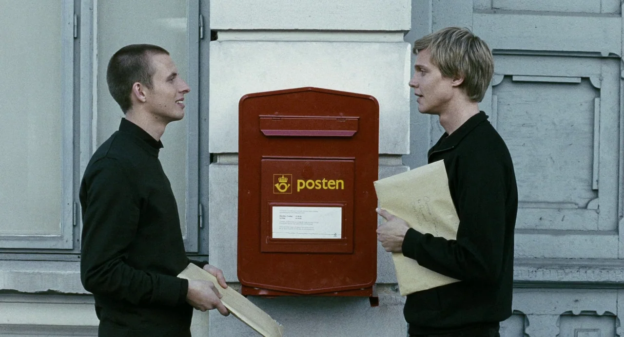 Reprise (2006) movie still: medium — Two men stand on a sidewalk against a pale gray building facade with a mailbox. Each man holds a manila…