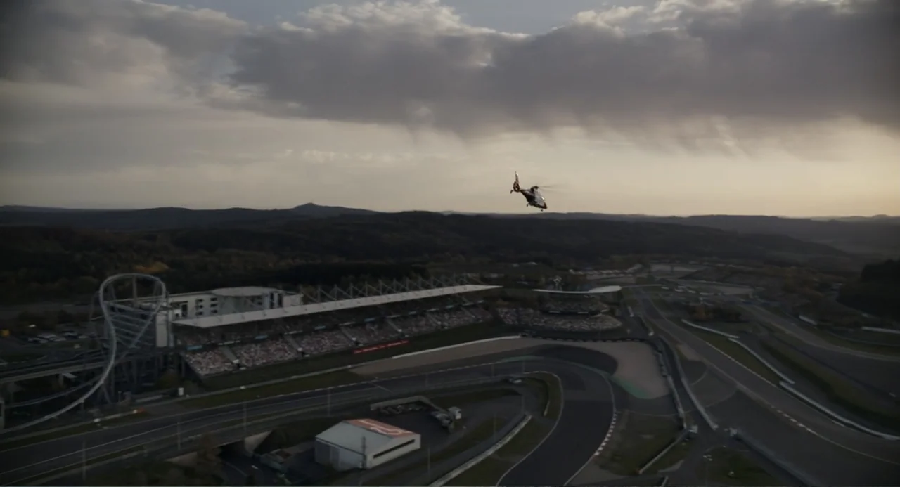 Gran Turismo (2023) movie still: establishing — A sprawling race track complex features grandstands, paved roads, and lush rolling hills. A small…