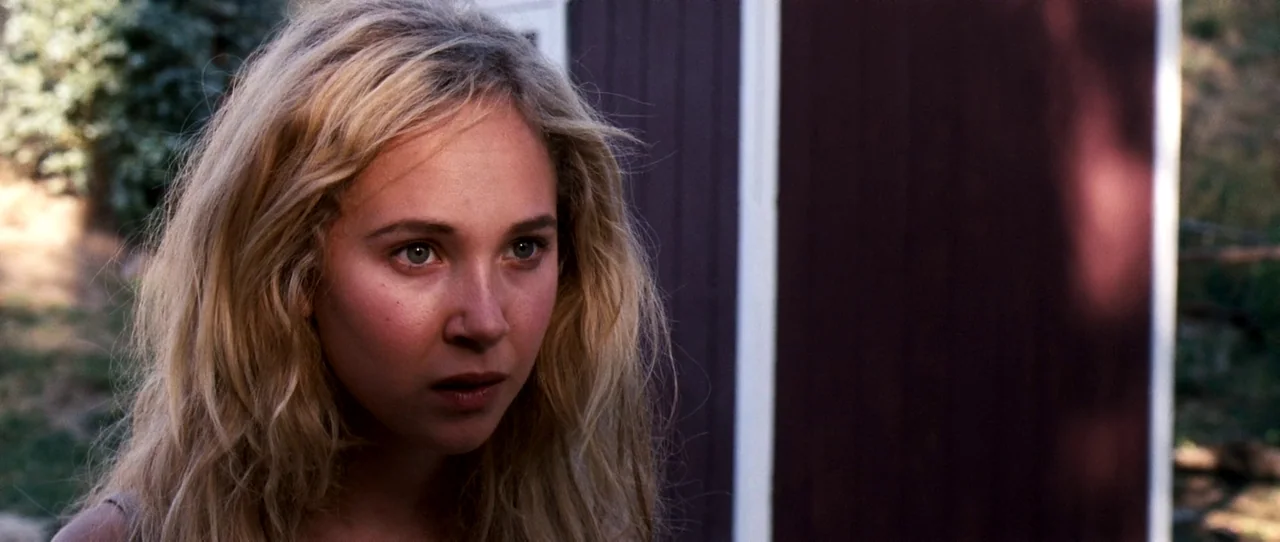 Little Birds (2011) movie still: closeup — A woman stands in front of a dark brown shed wall in an outdoor area. The subject looks off-camera with a…
