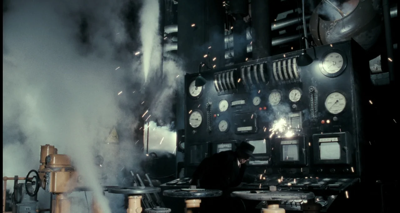 Batman (1989) movie still: wide — A dark, smoky industrial facility with pipes and a large vintage control console. A man in a dark hat and coat…