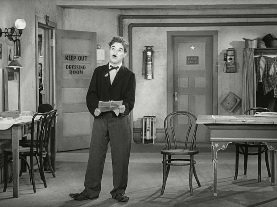 Modern Times (1936) movie still: fullbody — A utilitarian backstage area with wooden doors, a fire extinguisher, and simple furniture. A man in a…