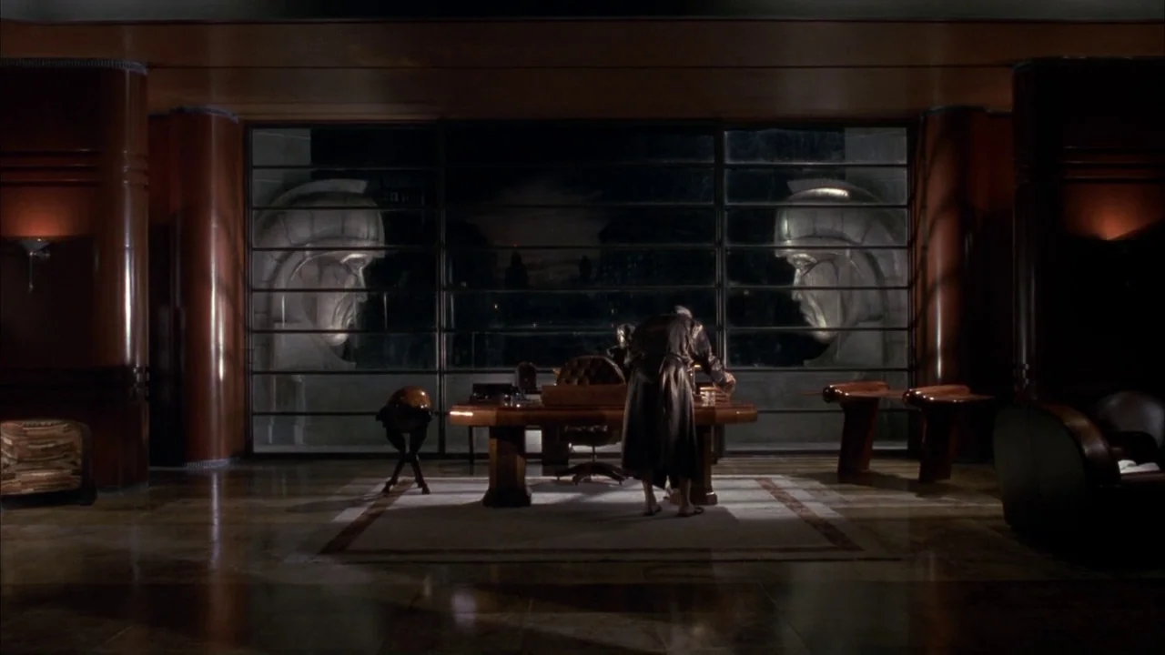 Batman (1989) movie still: wide — A dark, wood-paneled room features massive glass windows overlooking stone statues outside. A man stands alone at…
