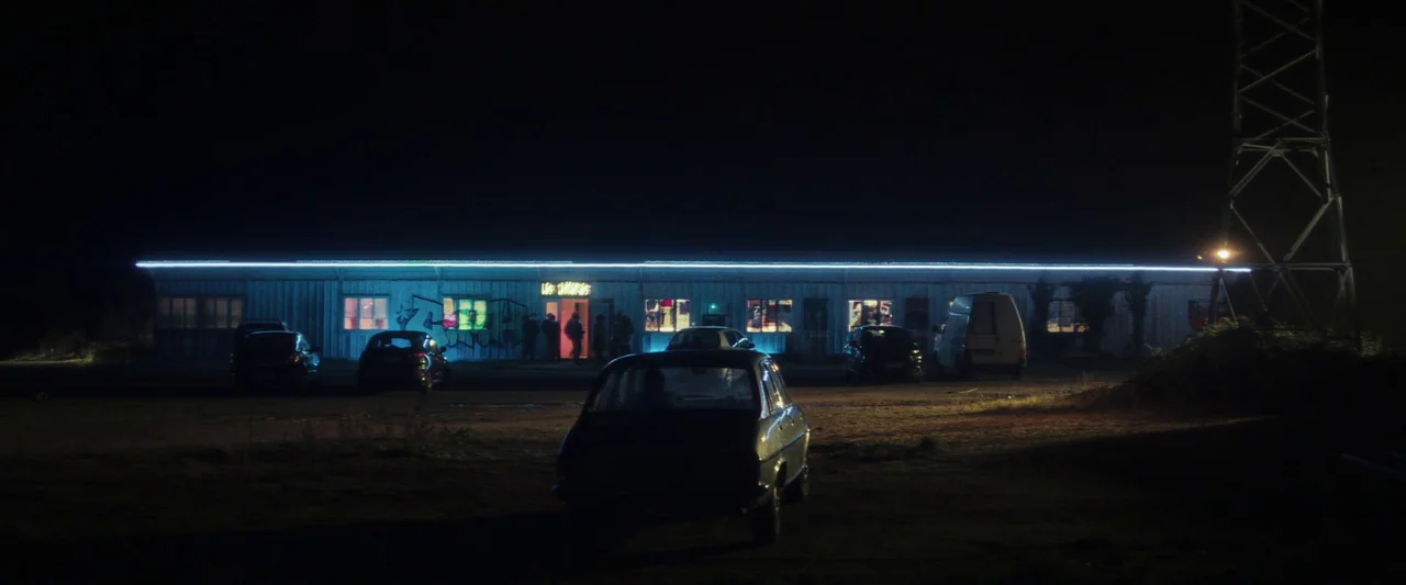 Vincent Must Die (2023) movie still: wide — A long, single-story industrial-style building sits in a dark, rural parking lot at night. Several…