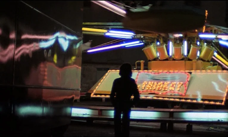 Small Change (1976) movie still: fullbody — A dark carnival at night features a spinning ride with illuminated yellow and blue lights. A lone figure…