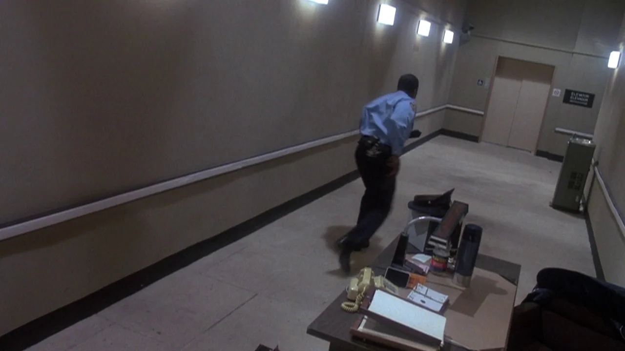 Re-Animator (1985) movie still: wide — A long beige hallway features a desk in the foreground and closed elevator doors. A security guard in a blue…