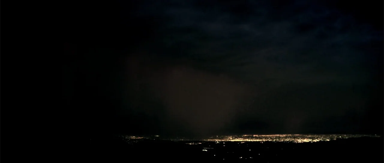 Rage (2014) movie still: establishing — A vast night horizon shows a distant sprawling city grid under a dark sky. The low-lying city lights glow…