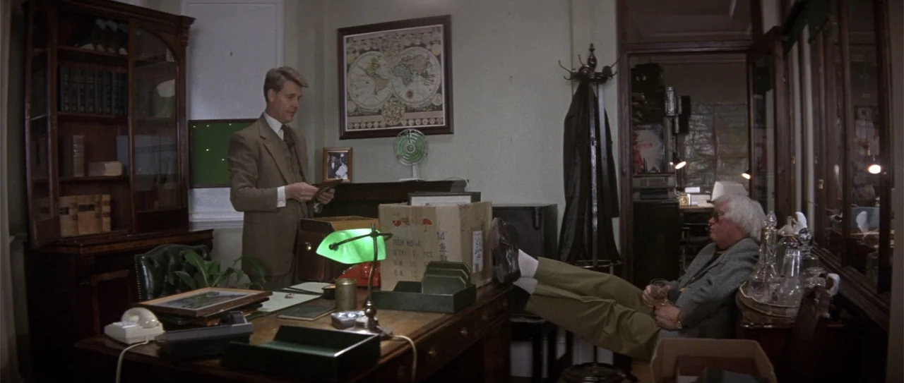 The Russia House (1990) movie still: medium — A cluttered 1980s office features dark wood furniture, a world map, and cardboard boxes. A standing…