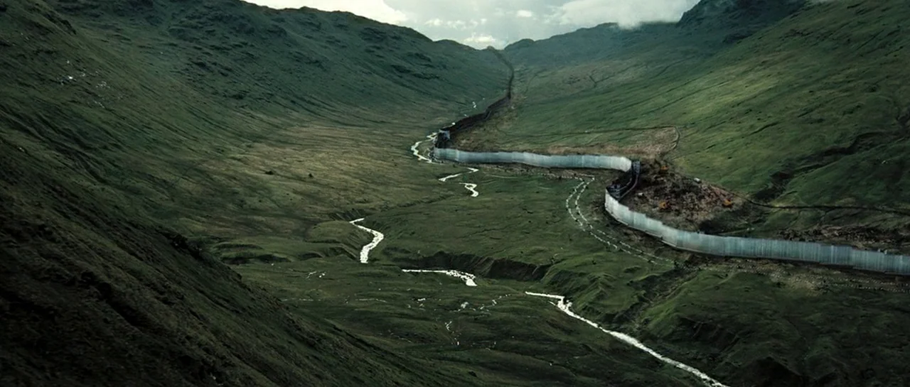 Doomsday (2008) movie still: establishing — A vast, green mountain valley is bisected by a long, concrete construction wall. A small river meanders…