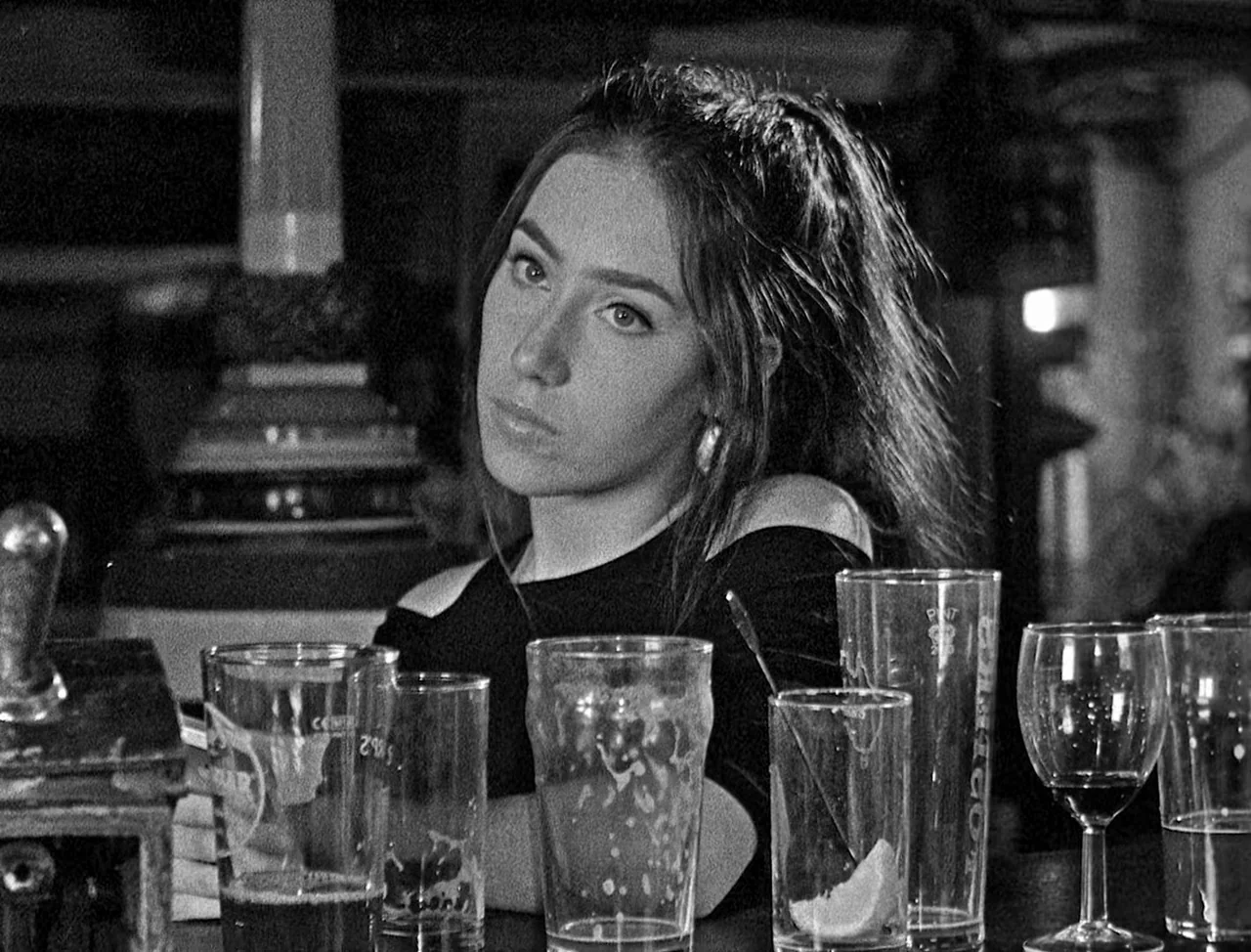 Bait (2019) movie still: medium — A dimly lit counter surface holds multiple empty glass cups and a wine glass. A woman sits behind the glasses with…