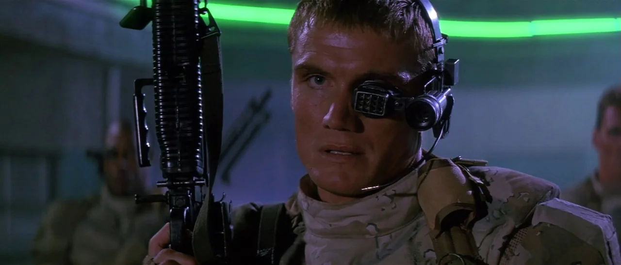 Universal Soldier (1992) movie still: medium — A dark military interior features low-key lighting and a glowing green neon tube background. A…