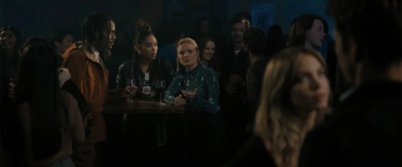 Anyone But You (2023) movie still: medium — A dimly lit tavern interior contains a group of young people at a table. Three people stand around a…