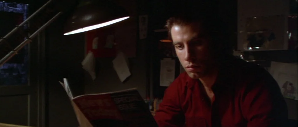 Blow Out (1981) movie still: medium — A dark room contains a desk with papers and a prominent articulated metal lamp. A young man sits in the dim…