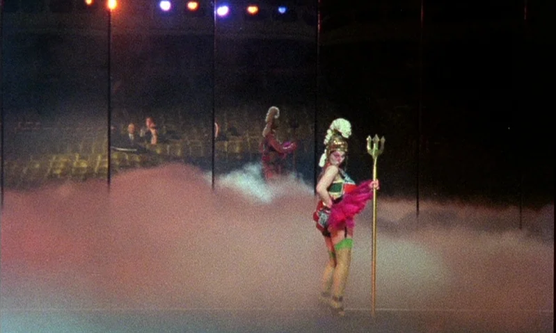 Jubilee (1978) movie still: fullbody — A performer in a theatrical costume stands on a stage obscured by low-lying smoke. Rows of empty auditorium…