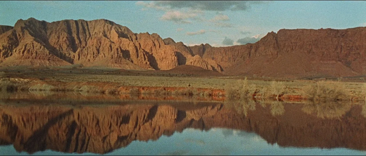 The Appaloosa (1966) movie still: establishing — A vast, arid desert landscape features red rocky cliffs reflected in a calm lake. Low-angle…