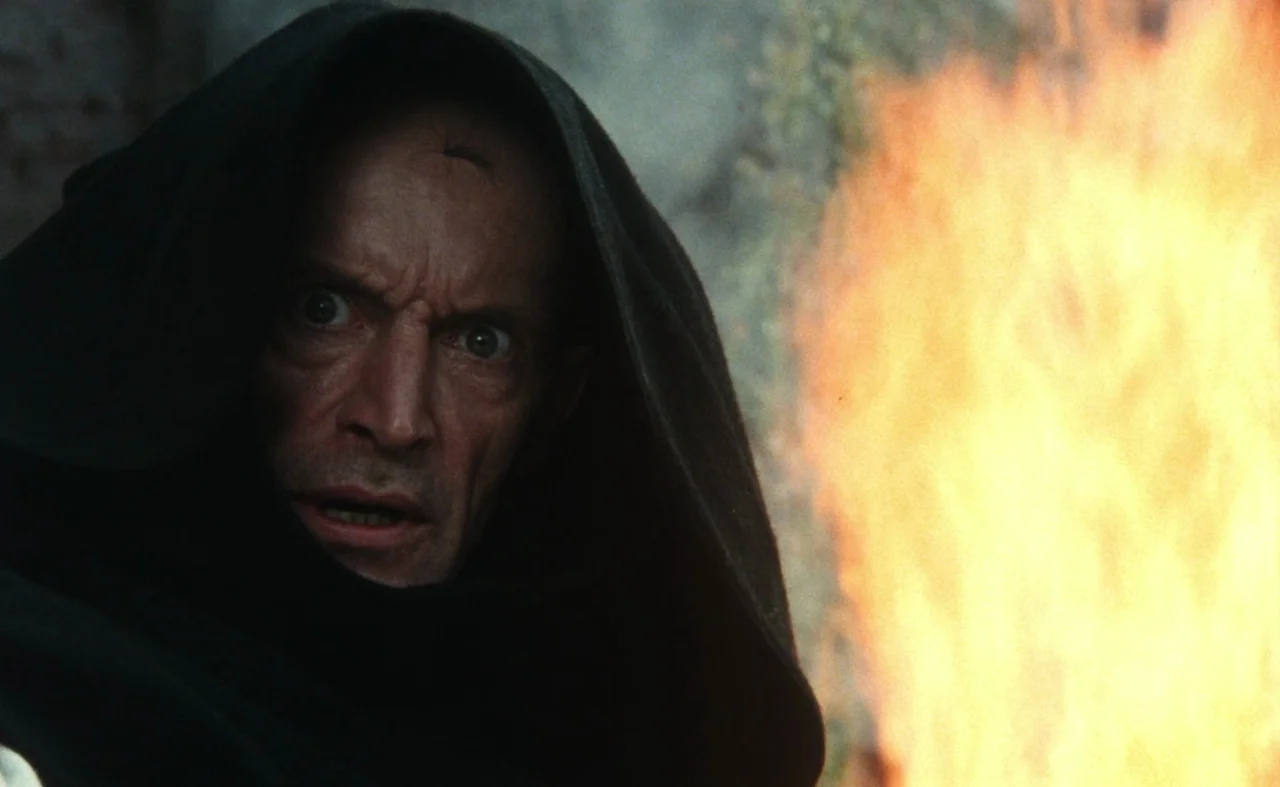 The Pit and The Pendulum (1991) movie still: closeup — A man in a dark cloak stands beside a blazing outdoor fire at night. His face is illuminated…