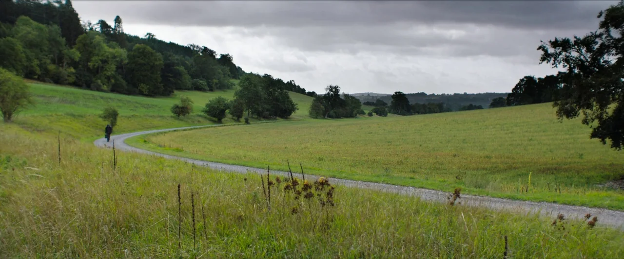 In The Earth (2021) movie still: wide — A rural gravel path curves through rolling green fields under a heavy, cloudy sky. A single person walks…