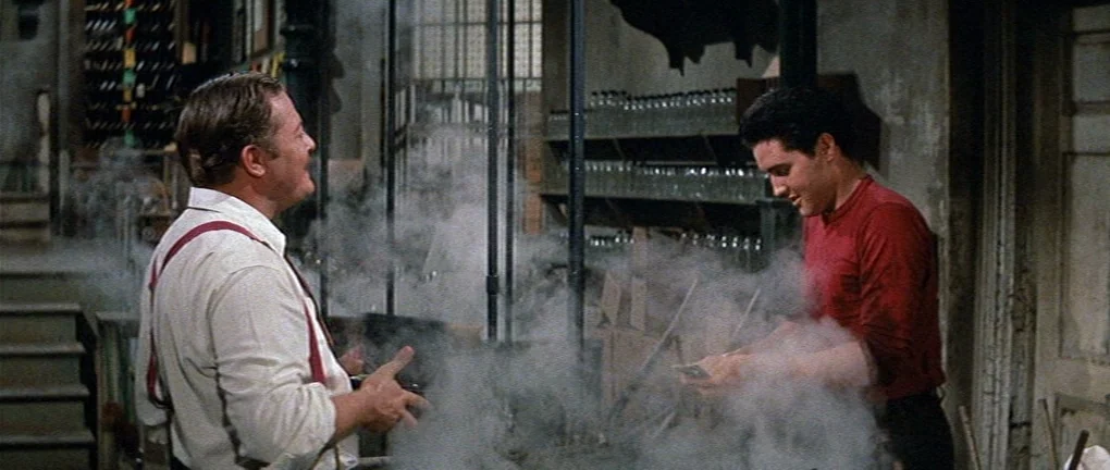 Wild in the Country (1961) movie still: medium — A dimly lit industrial basement contains shelves of glass bottles and heavy equipment. Two men…