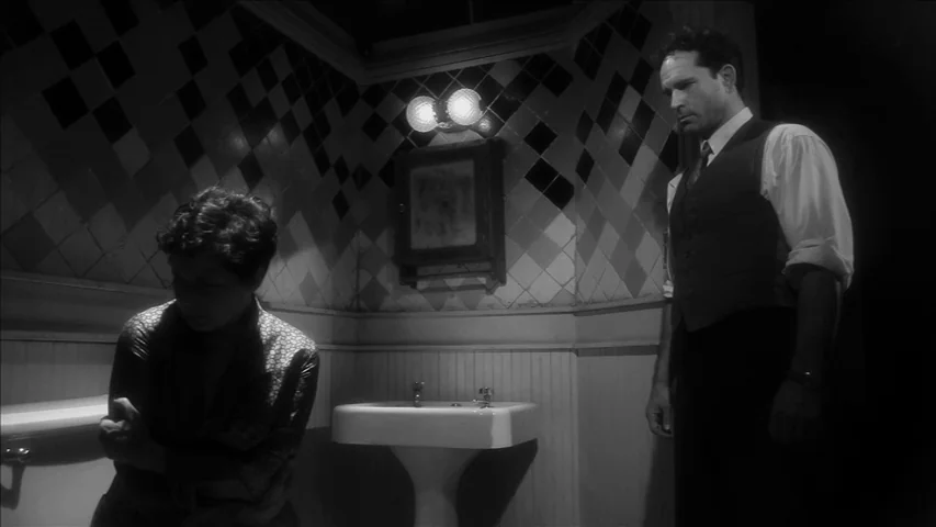 Keyhole (2011) movie still: medium — A cramped bathroom features diamond-patterned wall tiles and a pedestal sink under two bulbs. A man stands over…