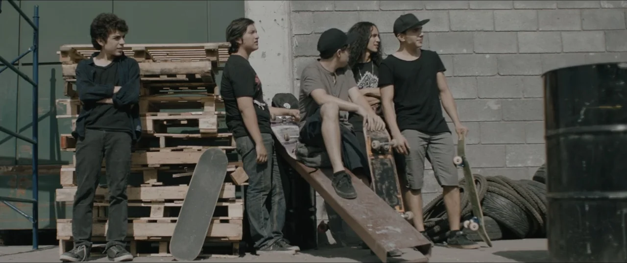 Death to Summer (2021) movie still: fullbody — A group of five young men congregate outdoors next to wooden shipping pallets. The men stand and sit…