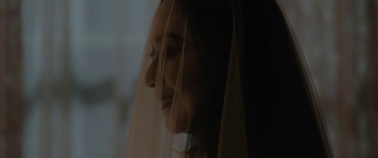 Satan’s Slaves (2017) movie still: closeup — A woman looks sideways through a fine mesh veil in a dim indoor setting. The veil partially covers her…